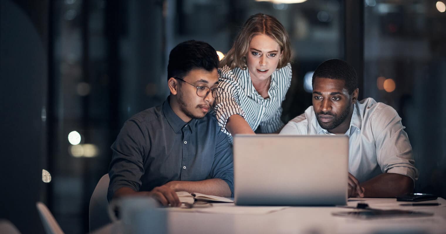 Three professionals collaborate late at night around a laptop, focused on AI Data Management, analyzing strategies for effective implementation.