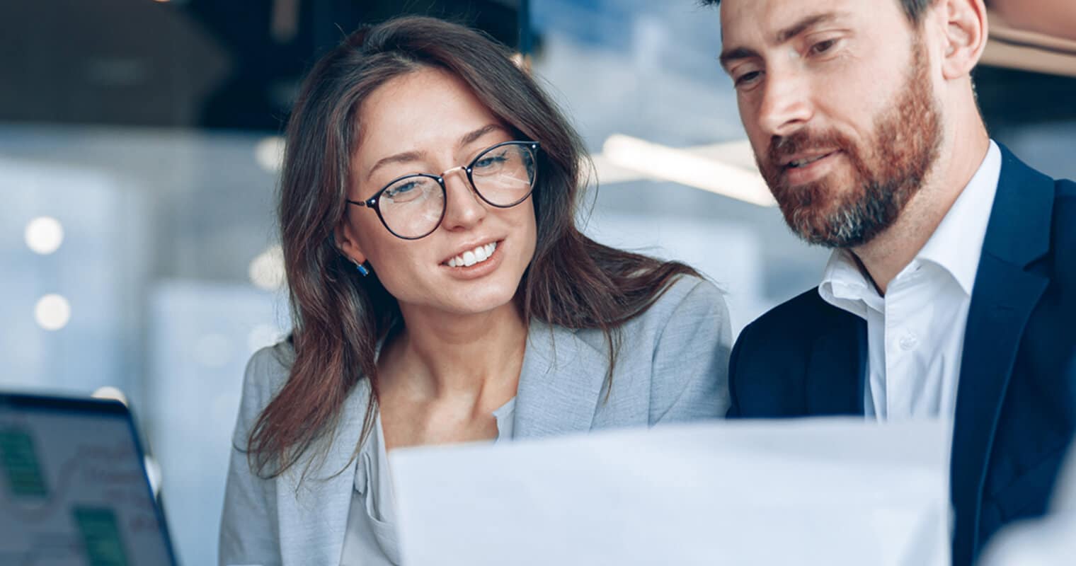 Two professionals, a man and a woman, in business attire are reviewing a document together in a modern office setting. The woman, wearing glasses, smiles as they discuss data management planning.